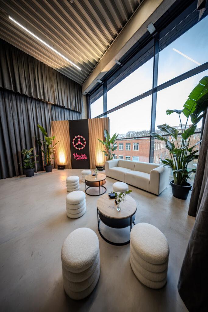 Eine moderne Lounge für Tech-Events mit weißem Sofa, gepolsterten Hockern, runden Tischen, großen Fensterflächen und zahlreichen grünen Pflanzen.